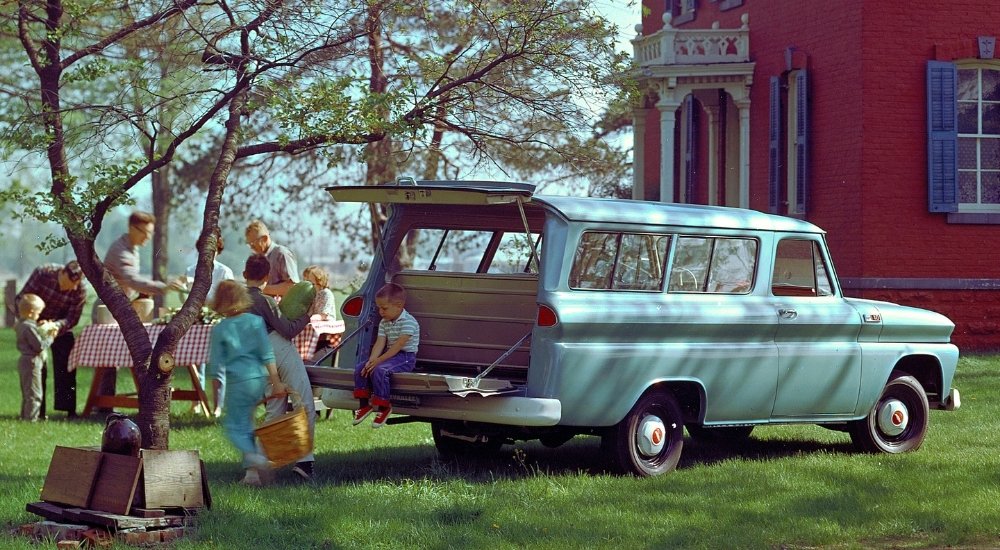 A baby blue 1965 Chevy Suburban parked with its trunk open at a backyard picnic.
