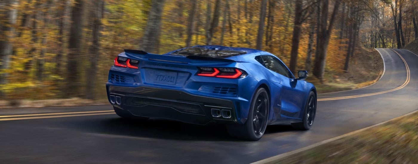 A blue 2025 Chevy Corvette E-Ray is shown driving on a tree-lined road.