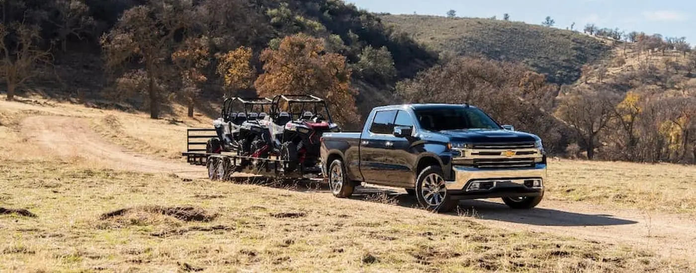 A black 2020 Chevy Silverado 1500 drives through a grassy field towing UTVs on a trailer.